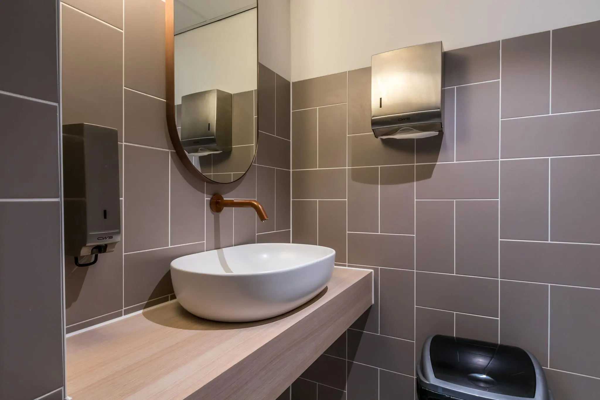 Modern wastafelmeubel met ronde spiegel en koperkleurige kraan in een betegelde toiletruimte aan de Vliegend Hertlaan.